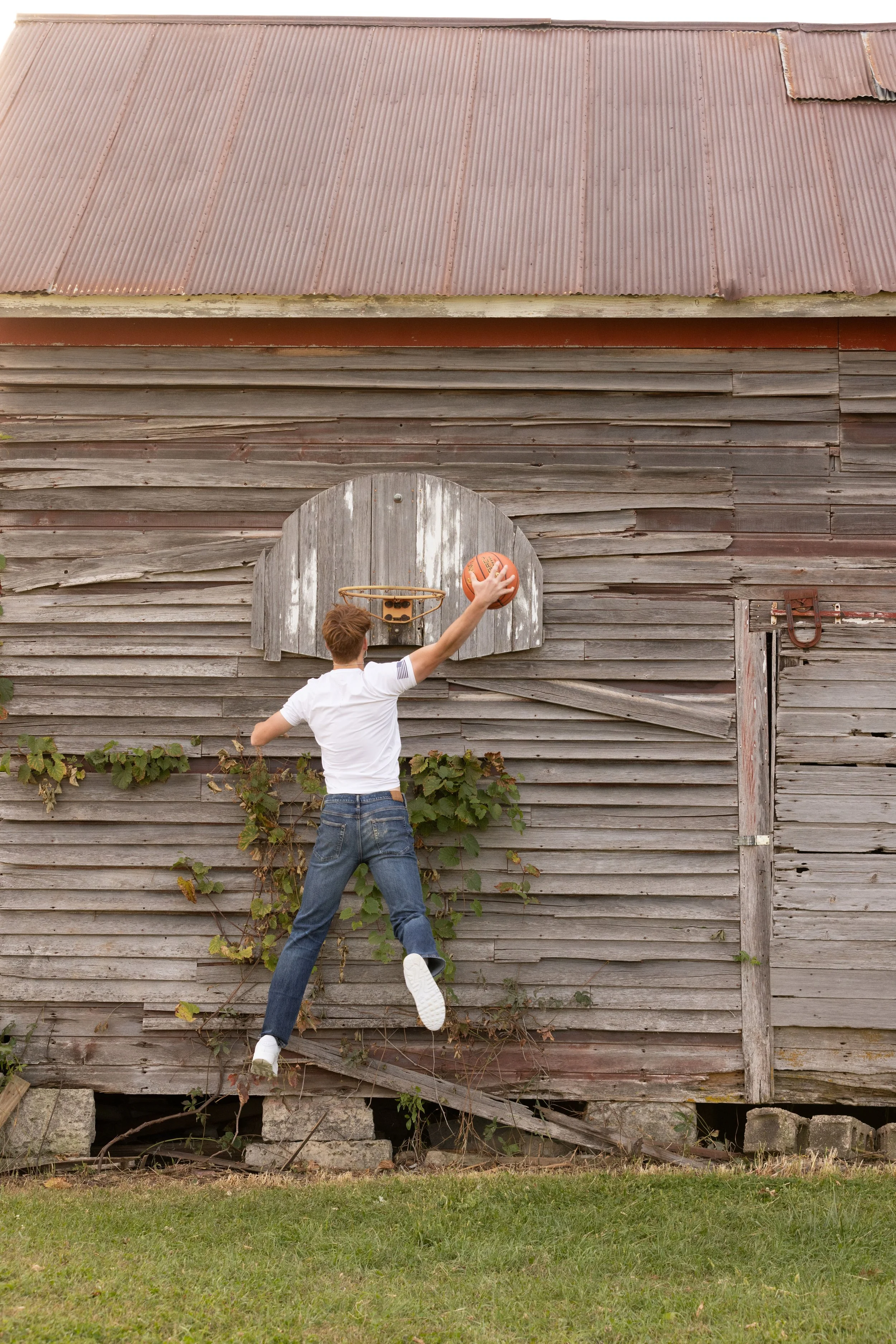 A young man in a white T-shirt and jeans playing basketball outdoors on an old wooden shed wall with peeling paint, jumping to make a shot.