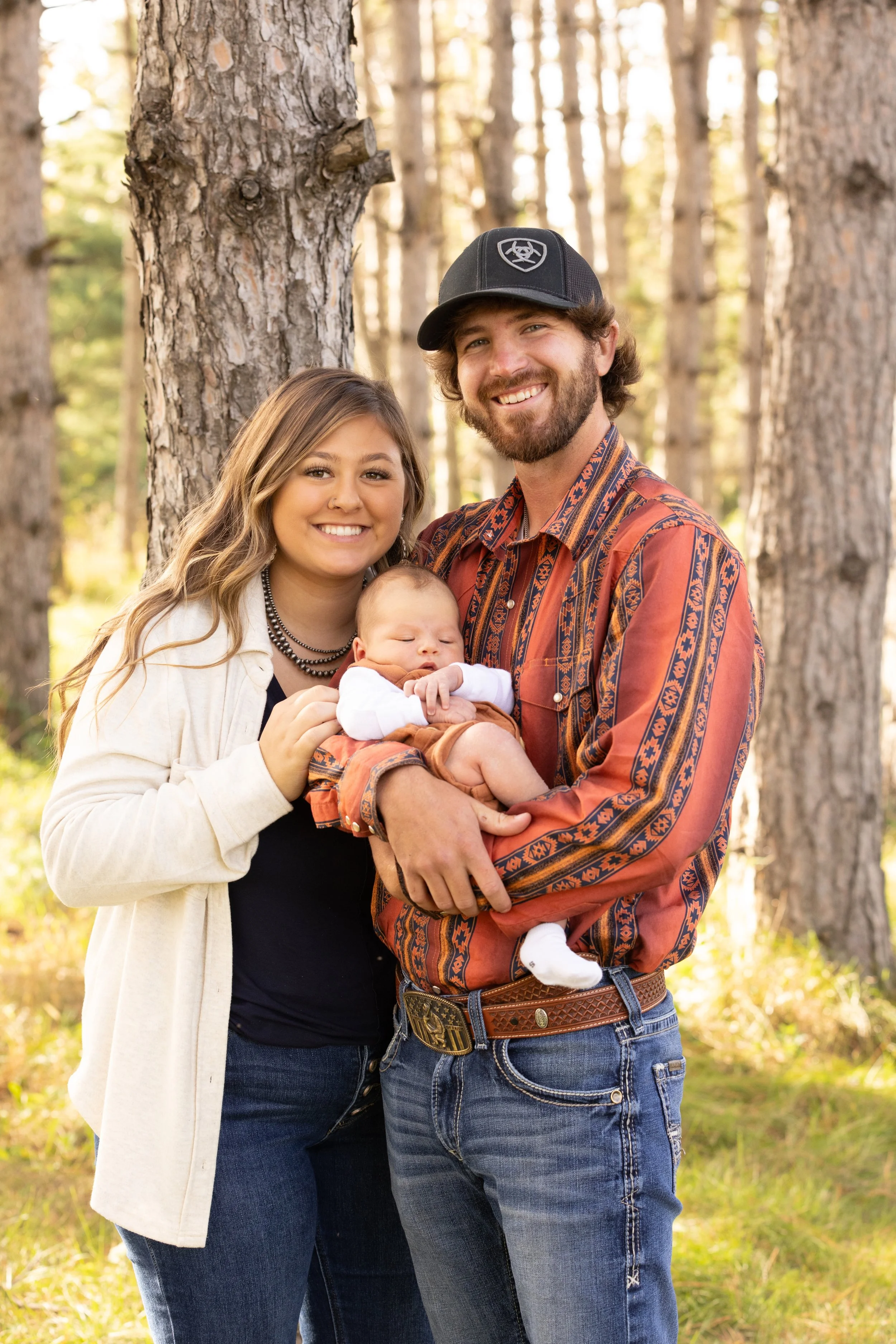 A young couple with their infant child in a forest, smiling at the camera with trees in the background.