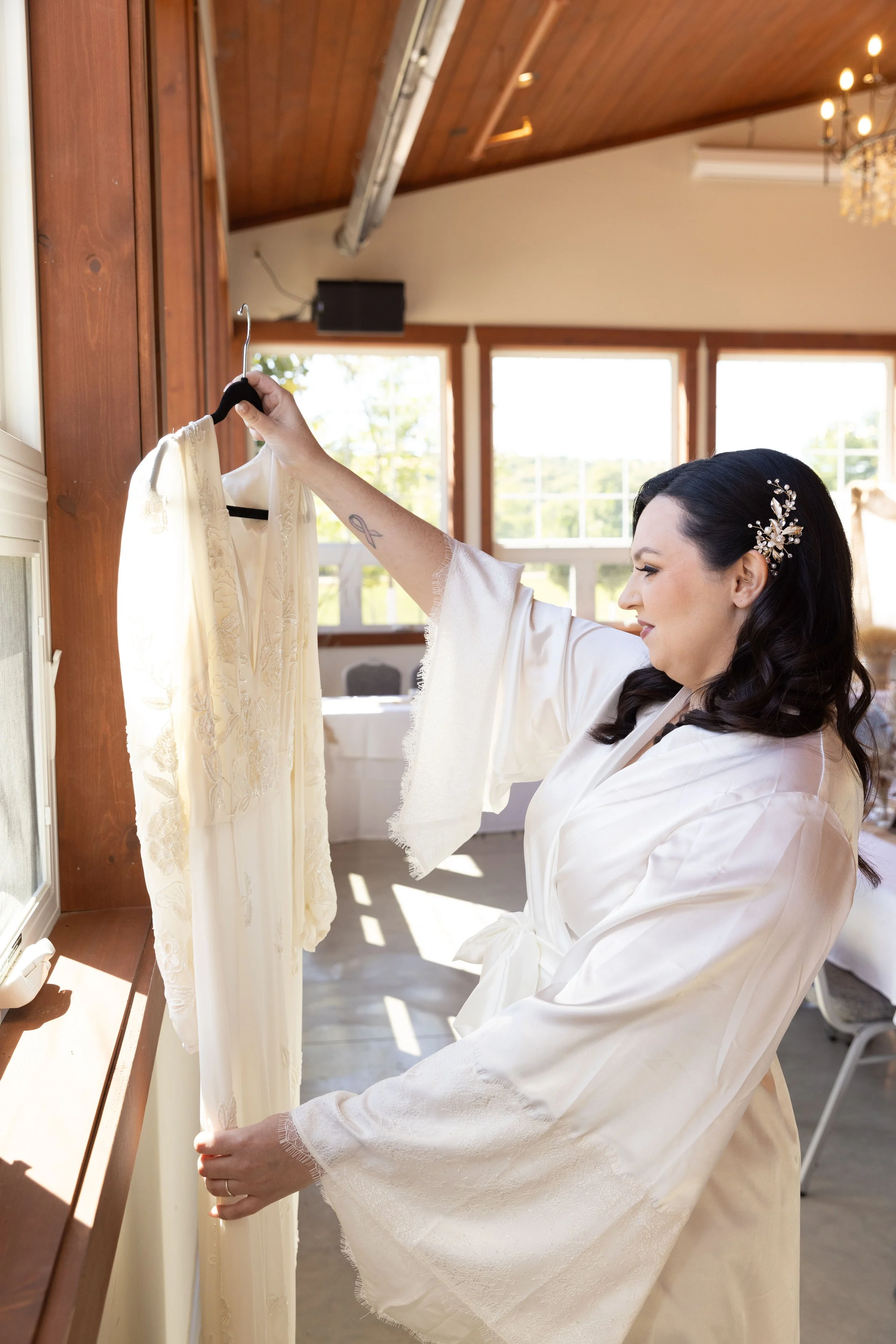A woman in a white satin robe with a hair accessory is smiling as she looks at a delicate cream-colored wedding dress with lace details, which she is holding up on a hanger in a sunlit room with wooden walls and large windows.