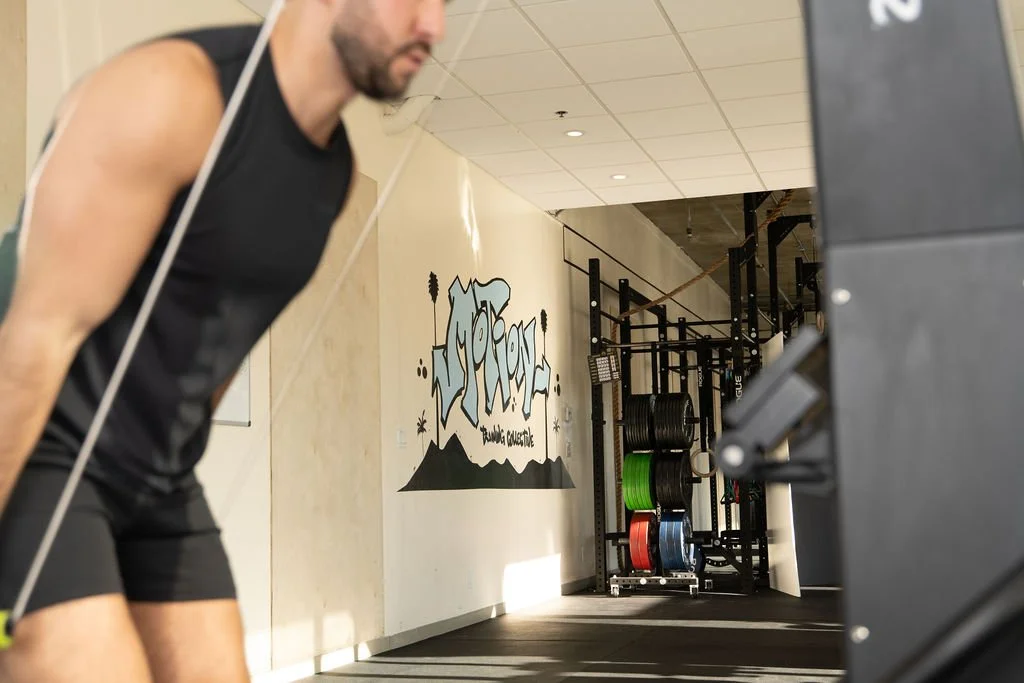 A man in workout clothes appears to be doing a gym exercise with a resistance band, with gym equipment and colorful weight plates visible in the background.
