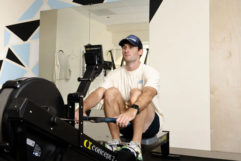 A man sitting on a rowing machine in a gym, wearing a white t-shirt, black shorts, a navy cap, and a smartwatch, looking focused.