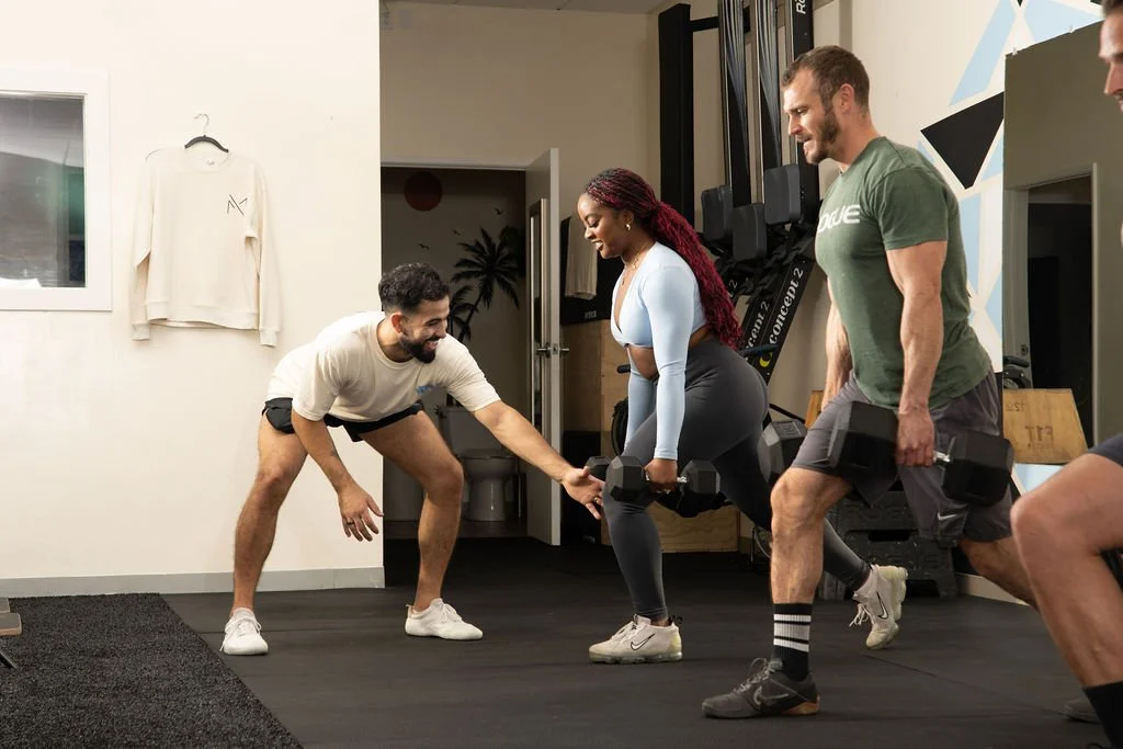 A fitness trainer assisting a woman in performing a squat with dumbbells in a gym, with another man standing nearby and a trainer in casual clothing guiding her.