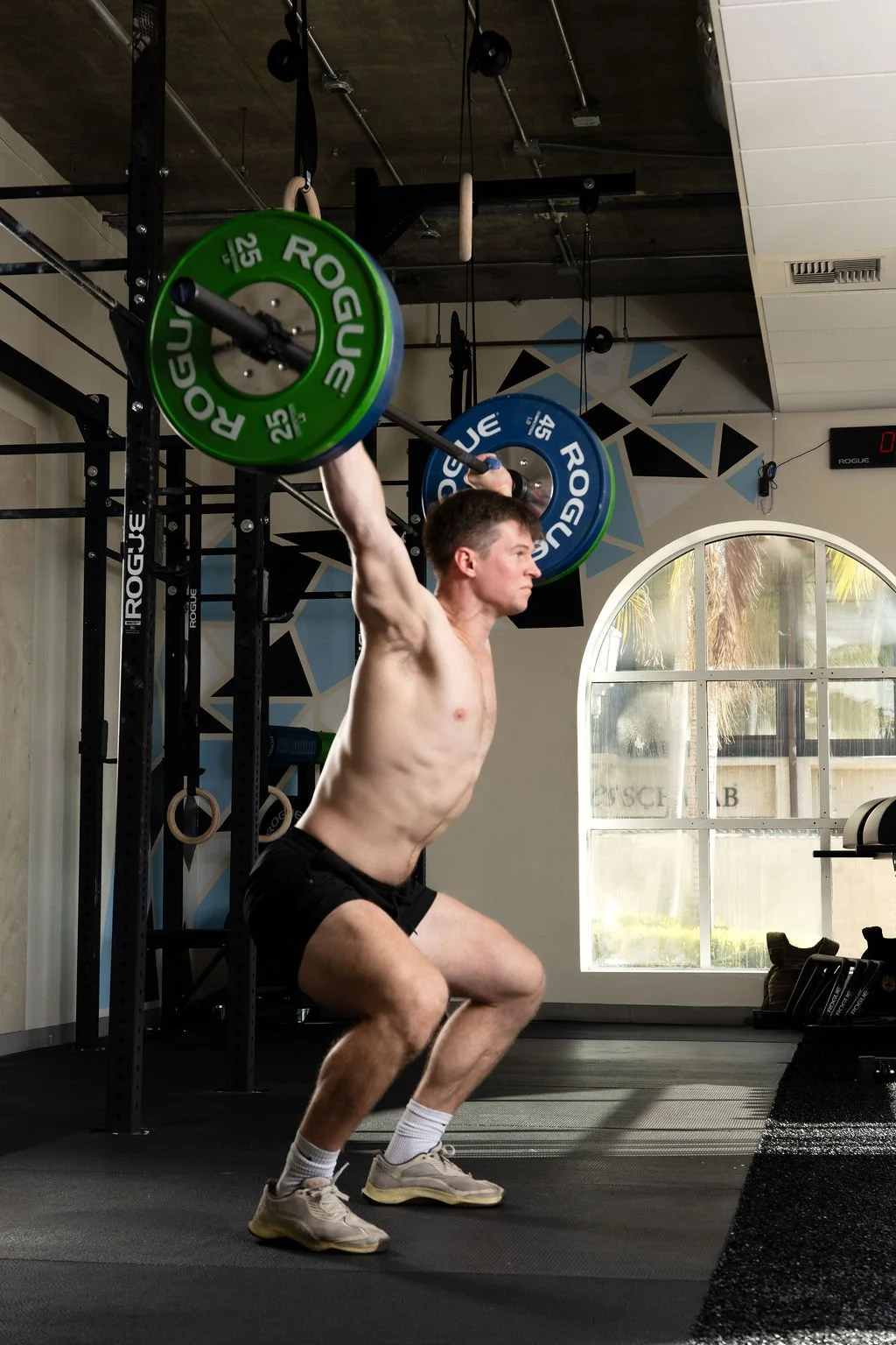 A man lifting a barbell with green and blue weight plates overhead in a gym.