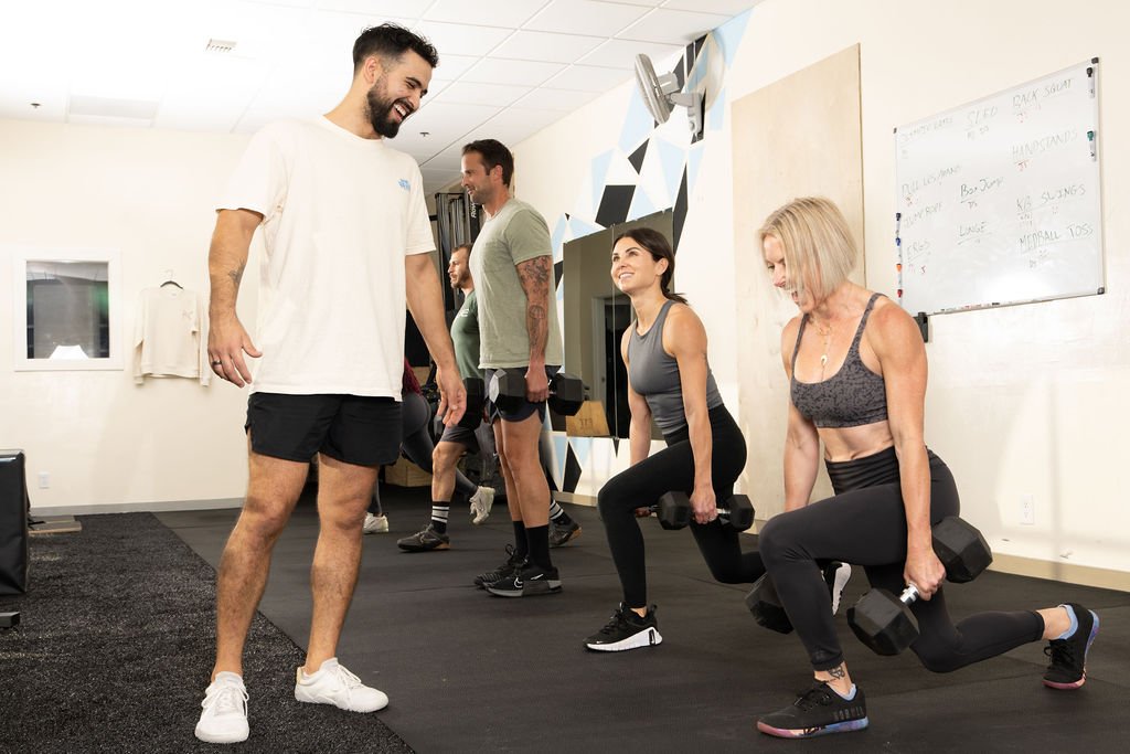 Six people working out in a gym, with three women using dumbbells in lunging positions and a man instructing them.