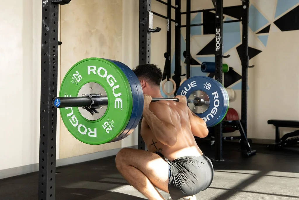 A muscular man with a tattoo on his side performing a squat with a barbell loaded with green, blue, and yellow weight plates in a gym.