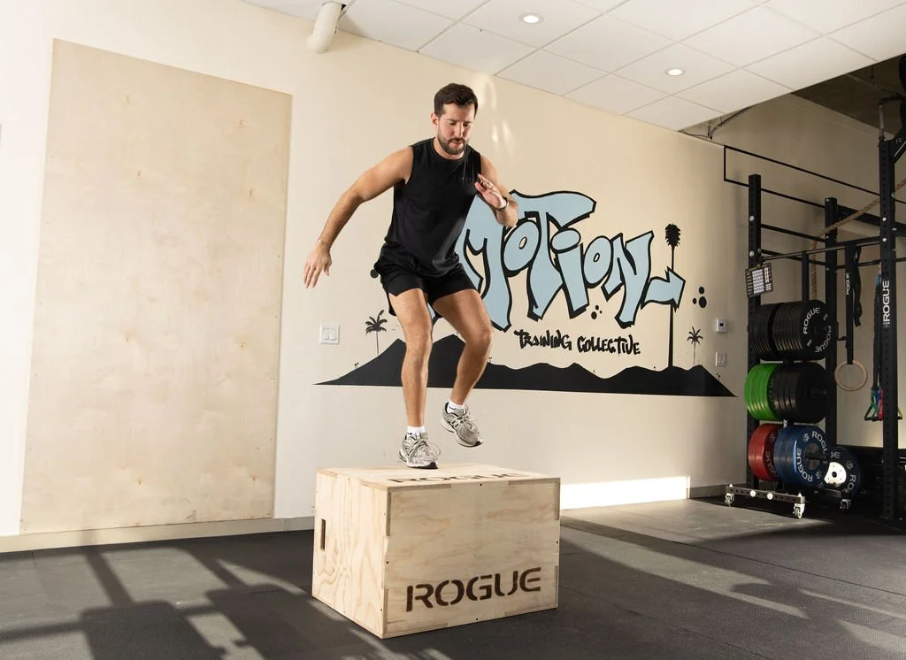Man doing Crossfit is jumping onto a plyometric box in a gym with a graffiti wall and weightlifting equipment.