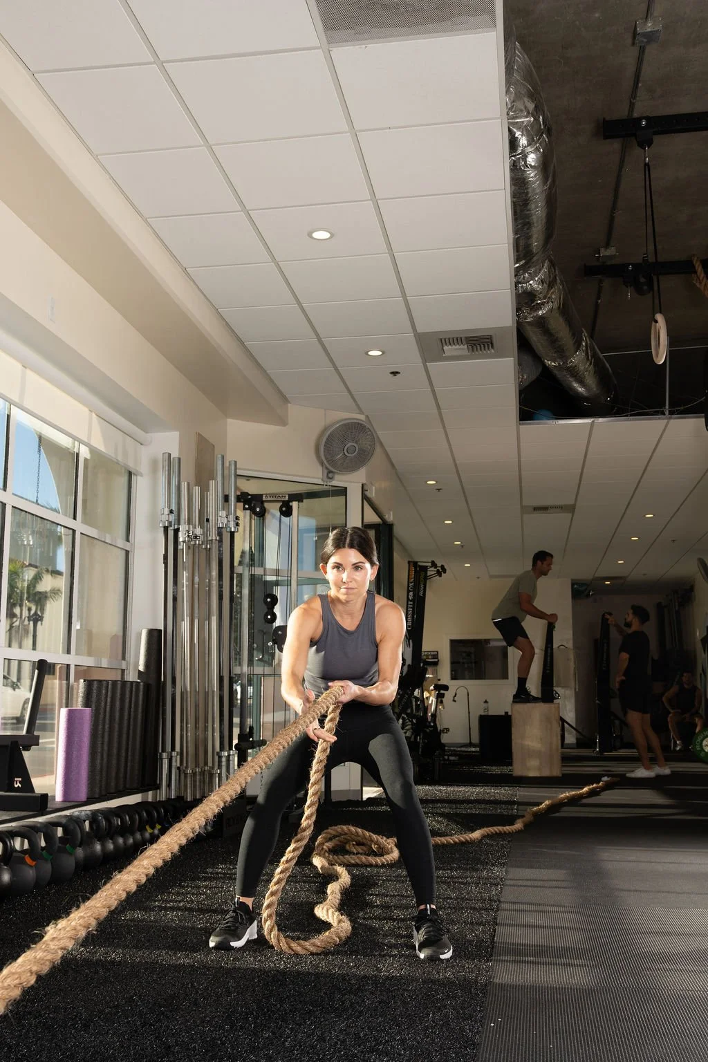 A woman in athletic clothing pulling a thick battle rope in a gym with other people working out in the background.