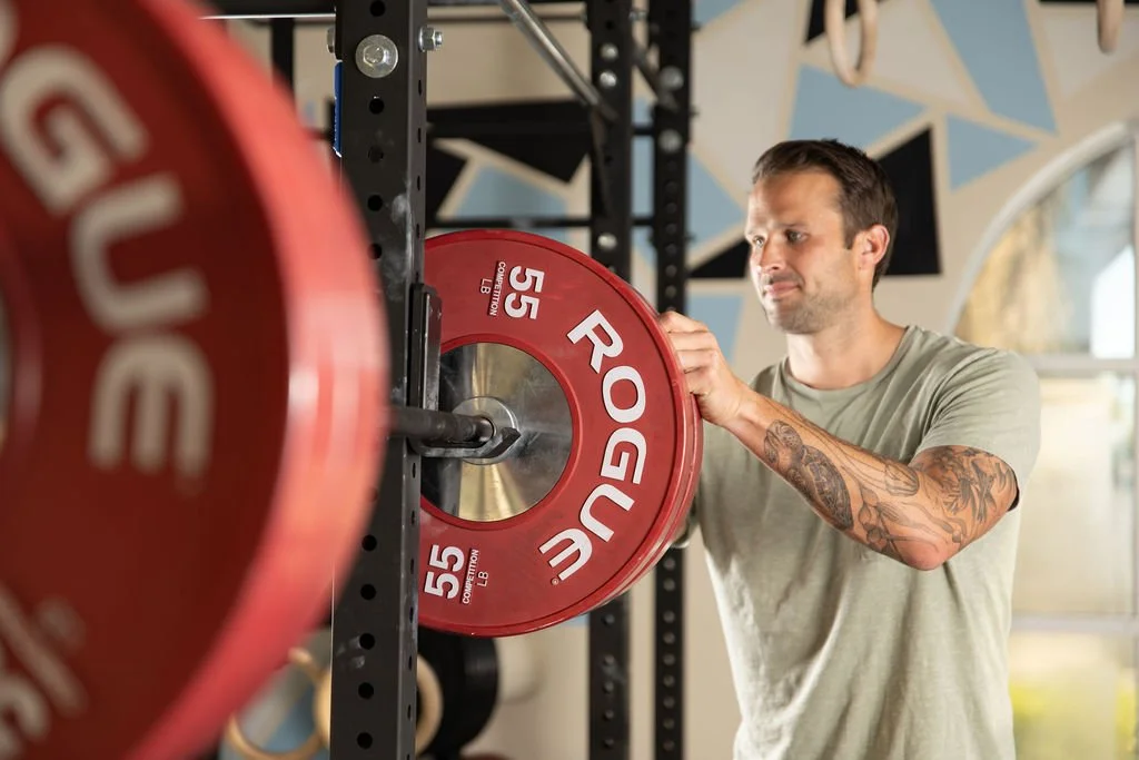 A man with tattoos on his right arm adjusting a red Rogue weight plate on a barbell in a gym.