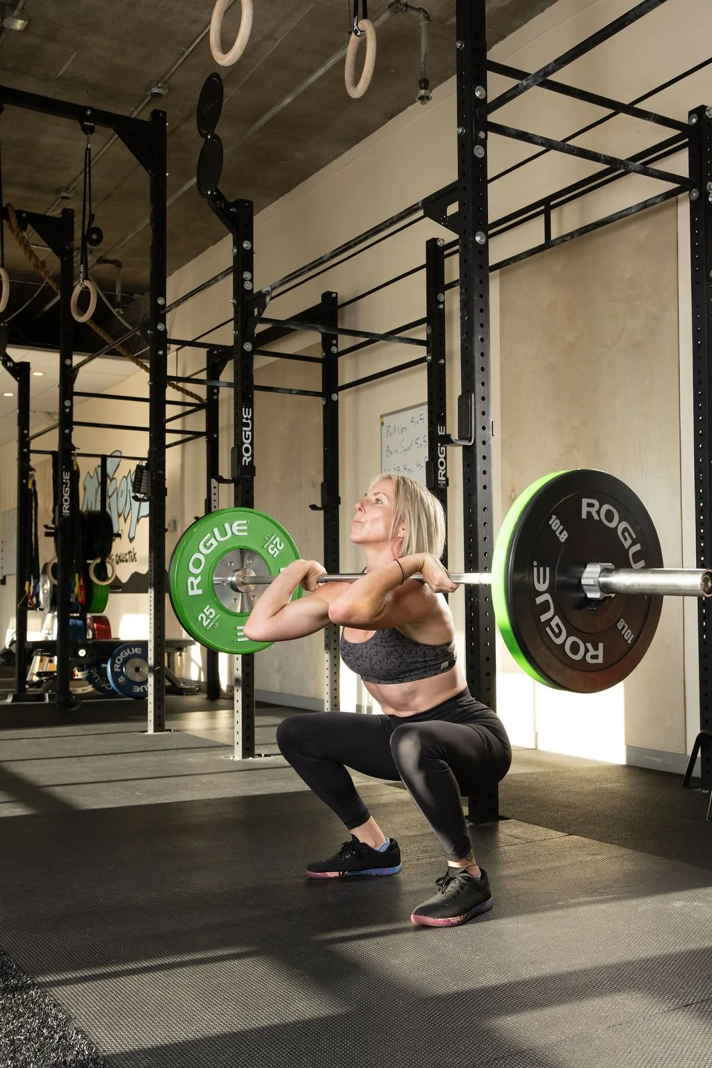 A woman doing a CrossFit workout wearing a sports bra and leggings performing a barbell squat in a gym. She is in a squat position holding a barbell with green weight plates on each side.