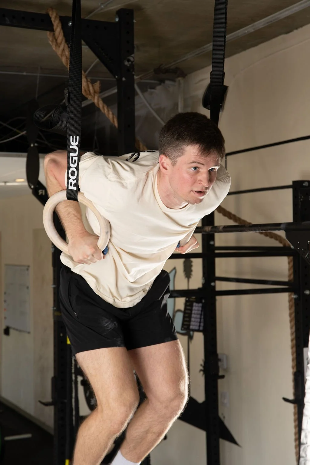 A man is doing a dip exercise on gymnastic rings in a gym, with a focused expression on his face, wearing a light-colored t-shirt and black shorts.  He is doing CrossFit.