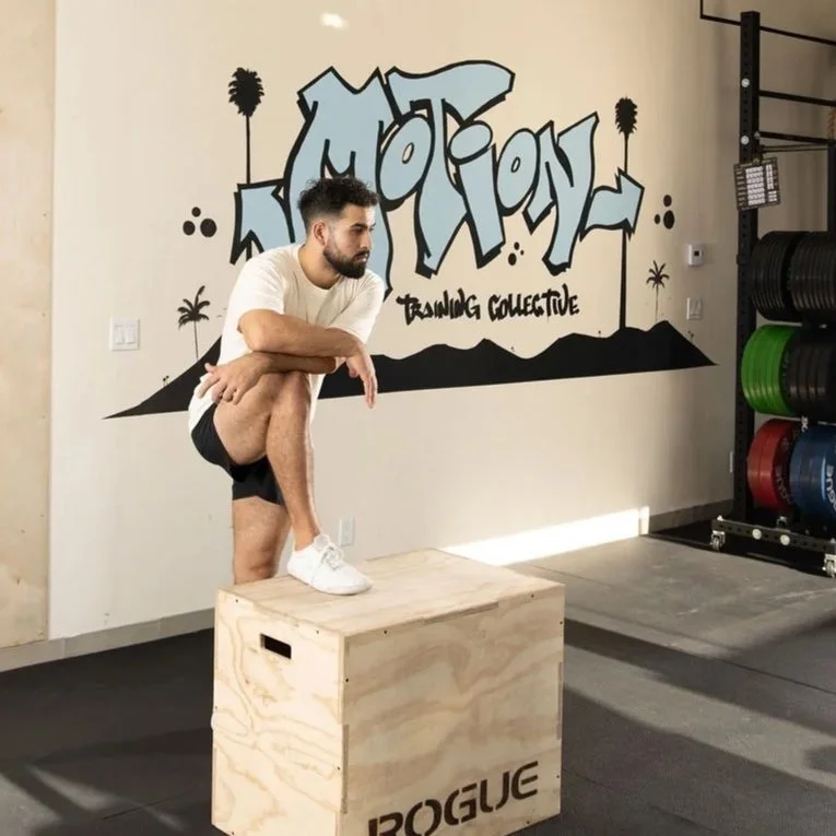 Man with beard in white t-shirt and black shorts performing a balance pose on one leg on a wooden plyometric box in a gym.