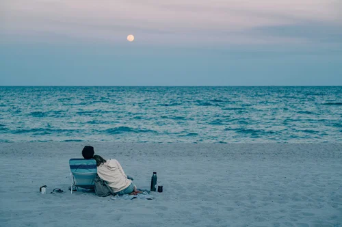 A couple sitting together on the sand watching the sea at dawn, the moon low in a soft sky, Miami Beach.