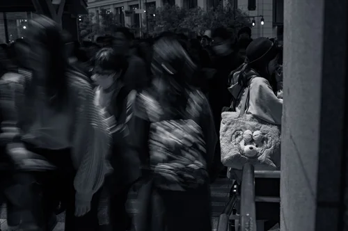 A blurred crowd hurrying past a still figure in kimono holding flowers on a busy street, Tokyo, in black and white.