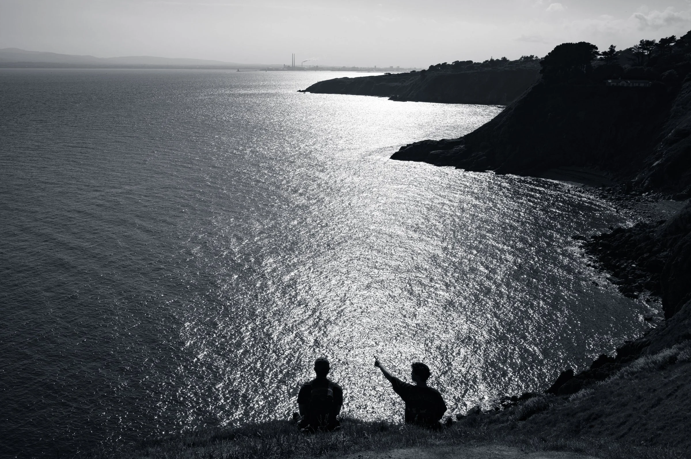 Two figures silhouetted on a clifftop looking out over a bay of glittering sunlit water, in black and white, a headland running into the distance.