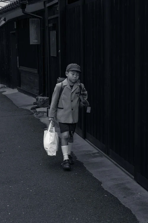 An older person walking down a narrow alley with a shopping bag, Kyoto, in black and white.