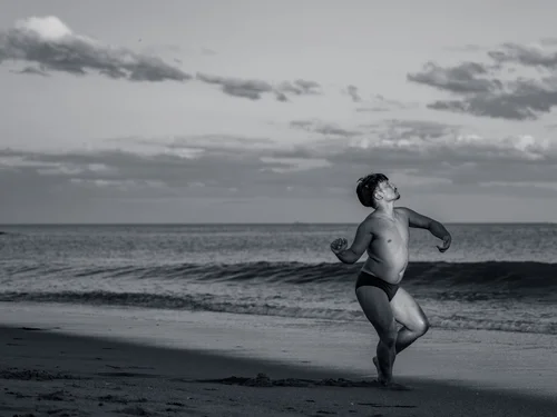 A dancer moving at the water's edge on the sand, waves and a cloudy sky behind at Rockaway Beach, New York, in black and white.