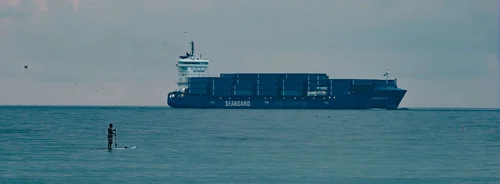 A large blue container ship on the horizon with a small paddleboarder in the foreground on calm water, Miami Beach.