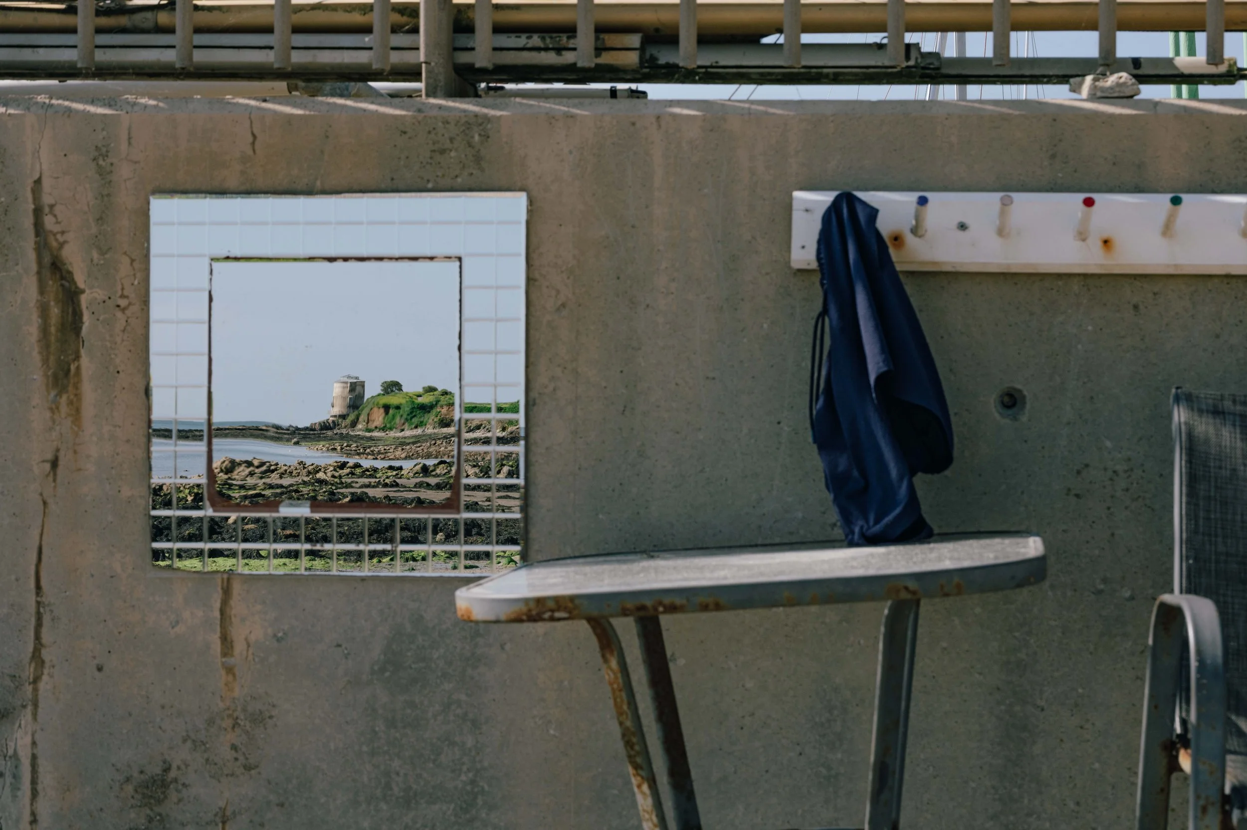 A seaside view framed through a square mirror mounted on a concrete wall, reflecting a Martello tower along the Dublin coast.