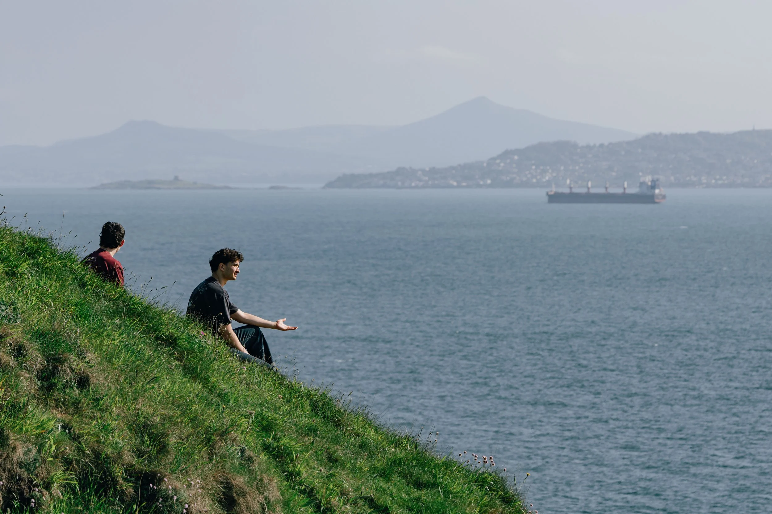 Two men sit on the green hillside above the sea, one gesturing mid-conversation, with Dublin Bay and a cargo ship in the background.