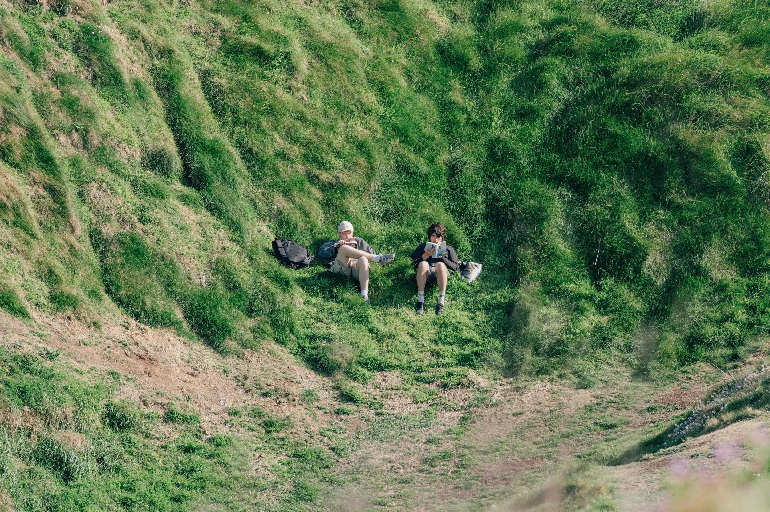 Two people lie back in a grassy hollow, reading and resting under soft afternoon light, framed by uneven cliffside grass.