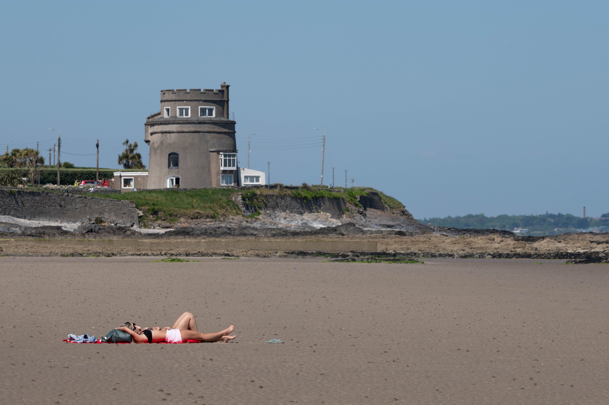A person sunbathes on a wide sandy beach beneath a Martello tower, the stone structure standing against a clear blue sky