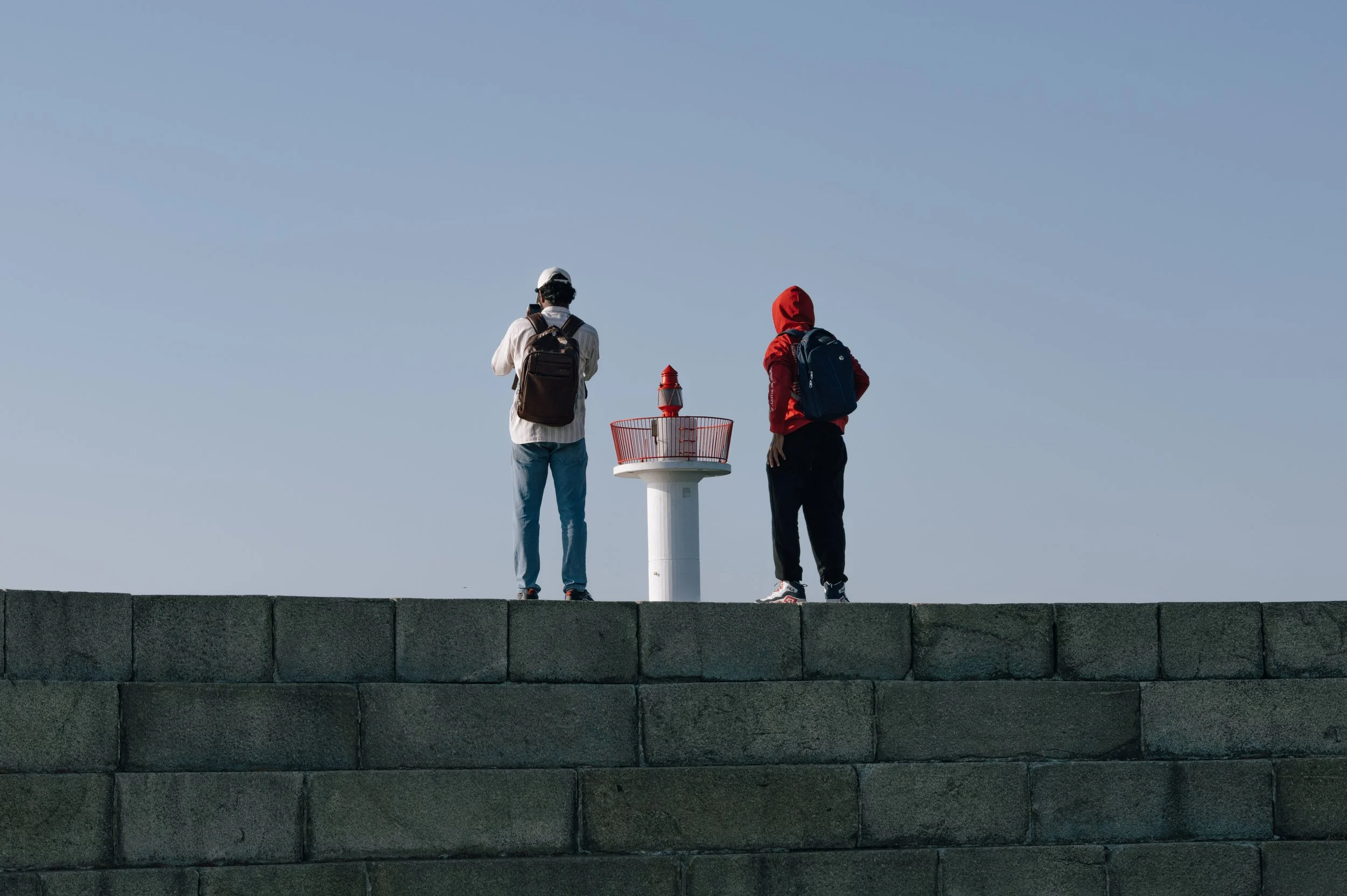 Two people with backpacks stand side by side on a pier, facing a small red navigation light against a clear blue sky.