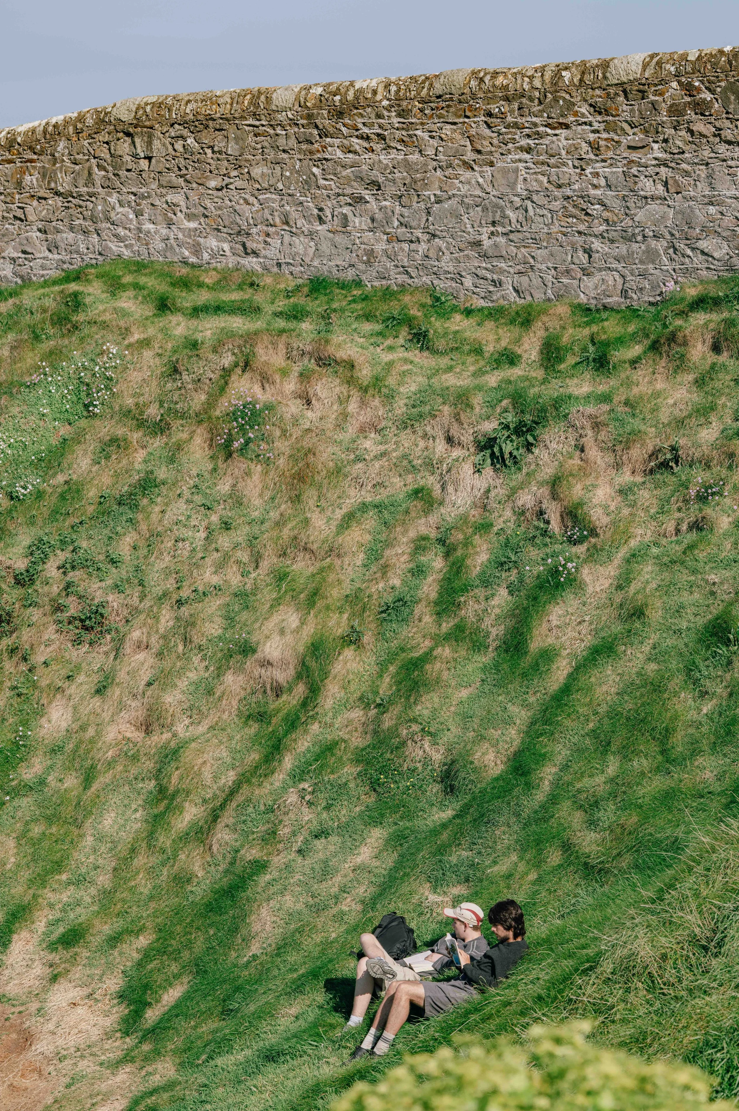Two people rest on the sloped grassy cliffs below a stone wall, the texture of the land patterned with brown and green patches.