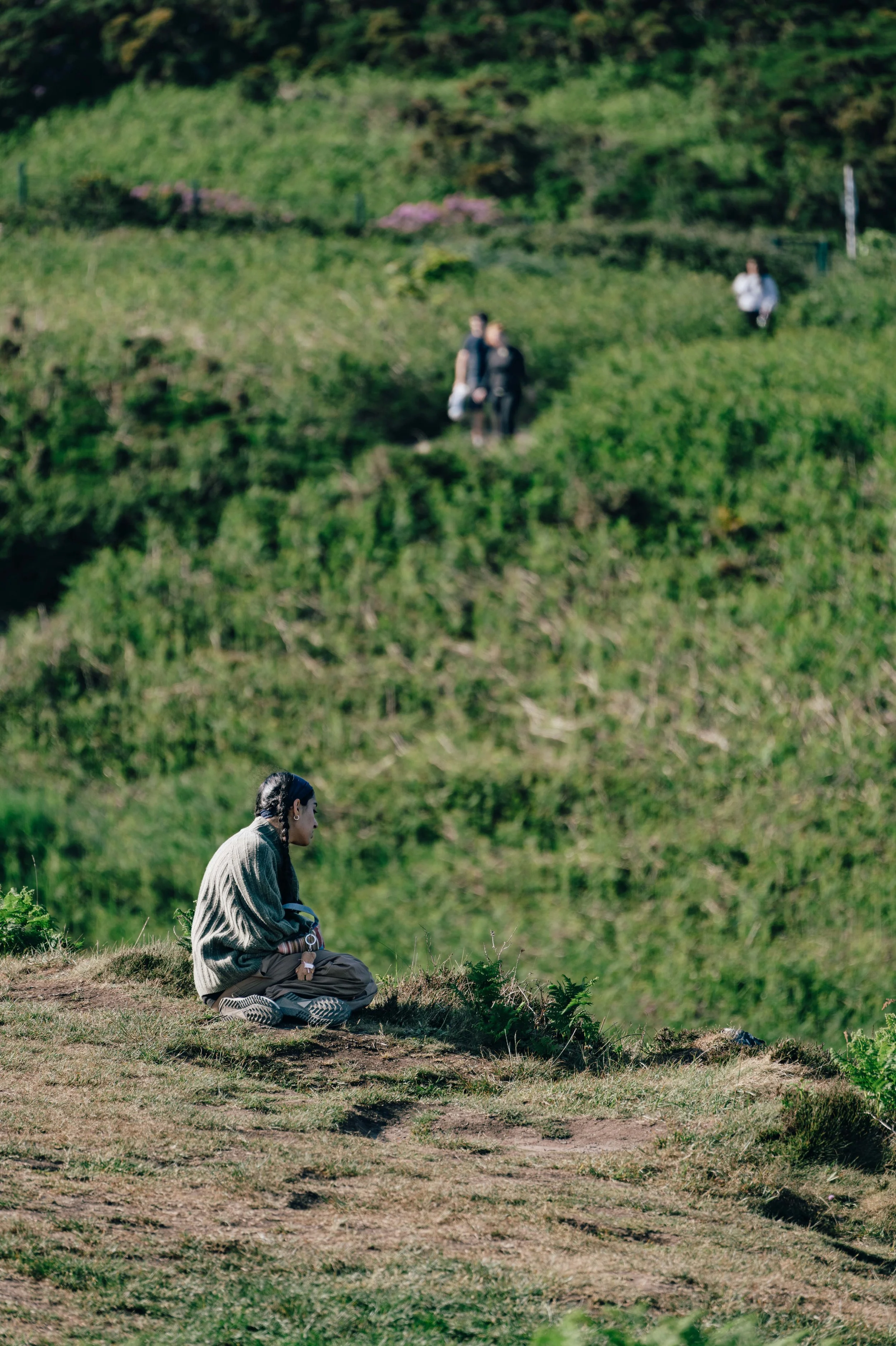 A solitary figure sits cross-legged on a coastal path, surrounded by green hills, gazing into the distance.