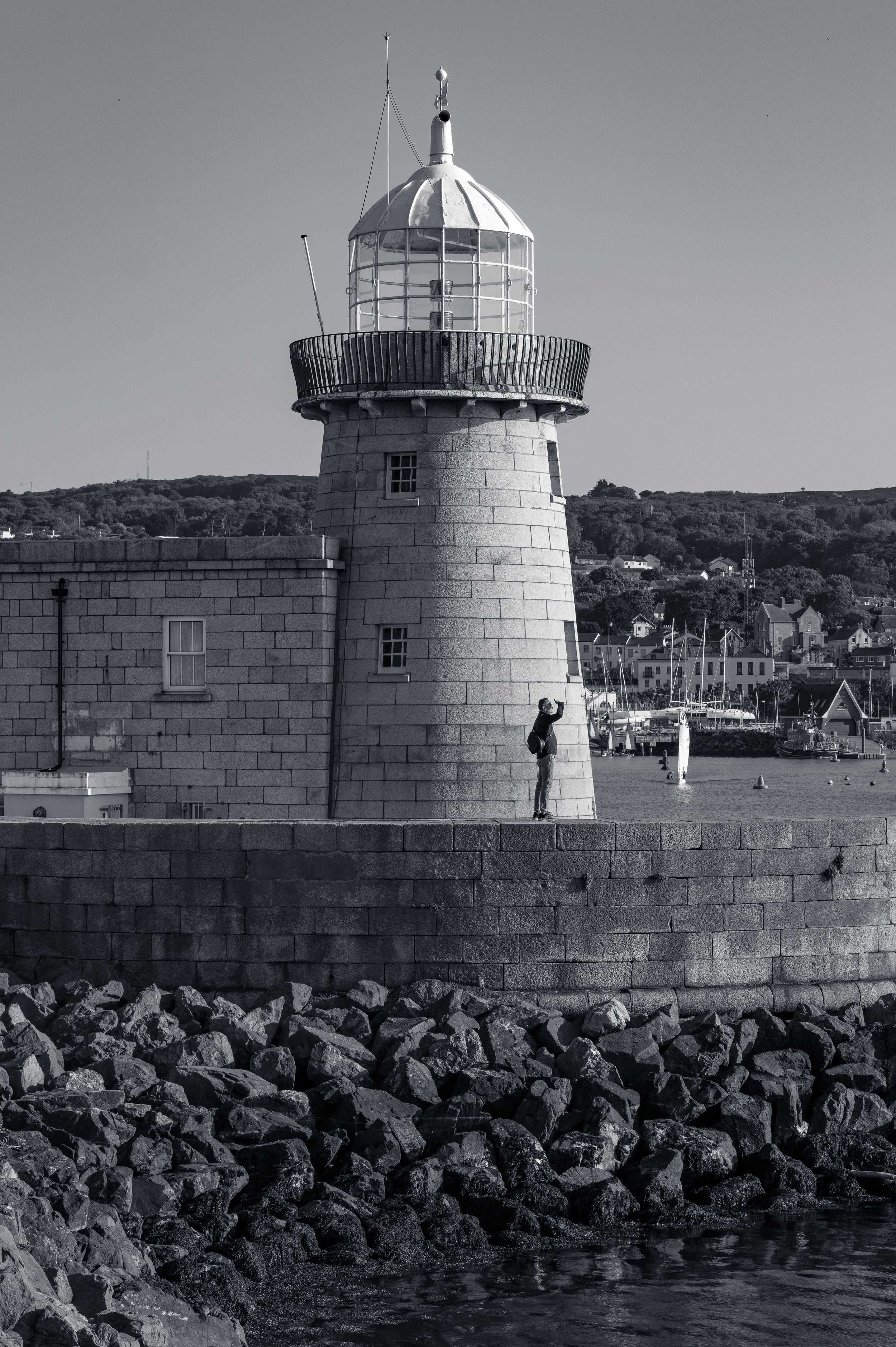 A person stands below the stone lighthouse at Howth Harbour, looking up toward its glass dome in the late afternoon light.