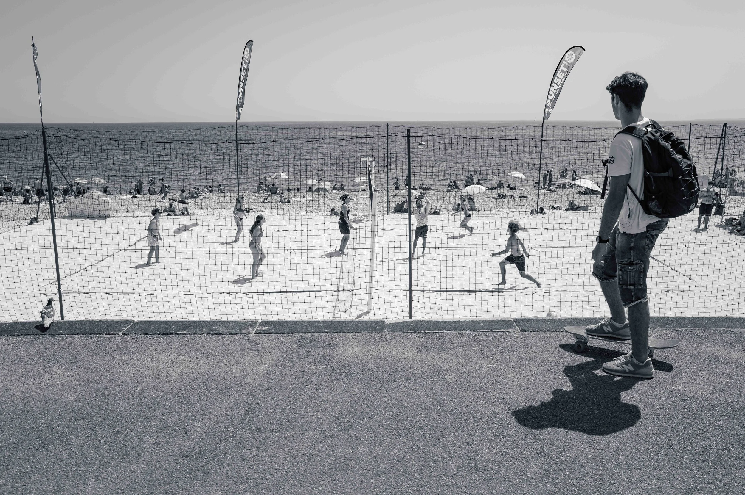 People playing beach volleyball on the sand beside the Promenade des Anglais, a passer-by watching from the walkway.