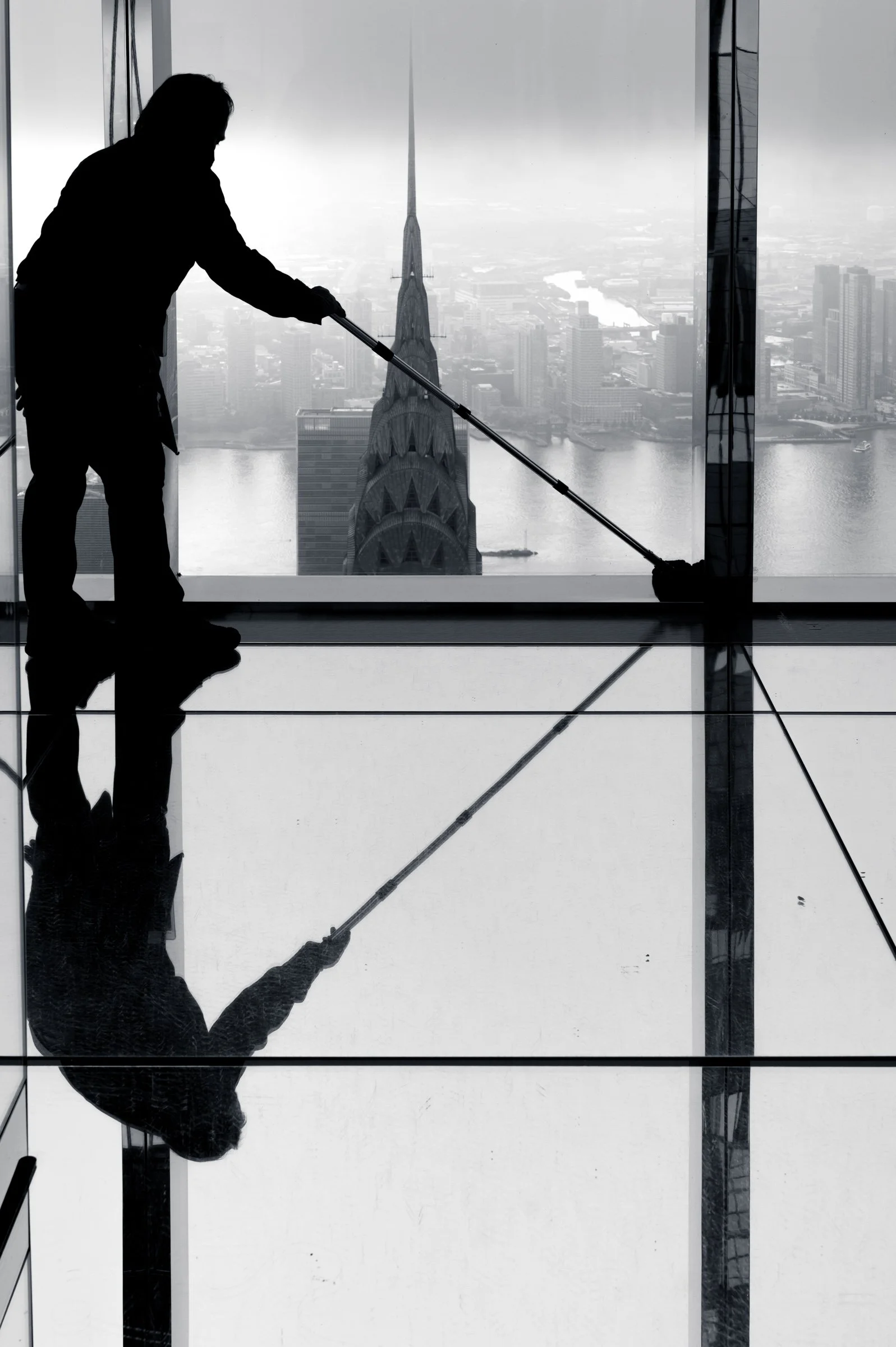 A cleaner on the glass at the top of Summit One Vanderbilt with a squeegee extended, the Chrysler Building spire directly behind her, New York, in black and white.