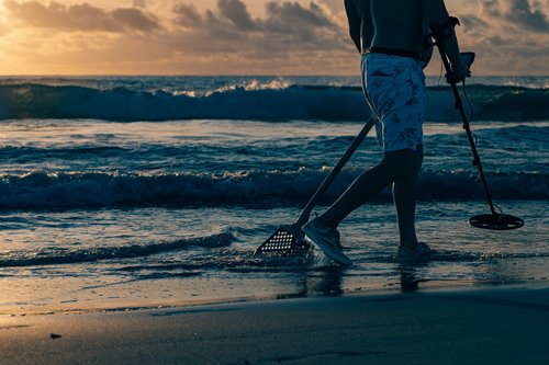A person sweeping the sand with a metal detector at the water's edge in warm light, Miami Beach.