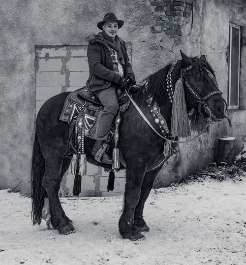 A rider in a hat and heavy coat mounted on a dark horse in the snow beside a building, Bucovina, Romania, in black and white.