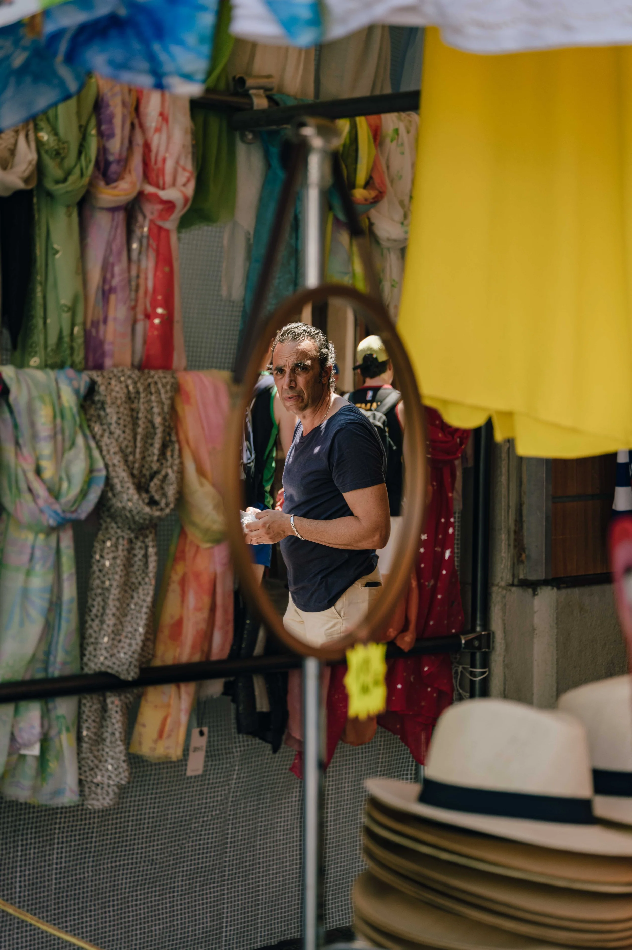 A man reflected in an oval mirror at a market stall hung with colourful scarves and Panama hats in Vieux Nice.