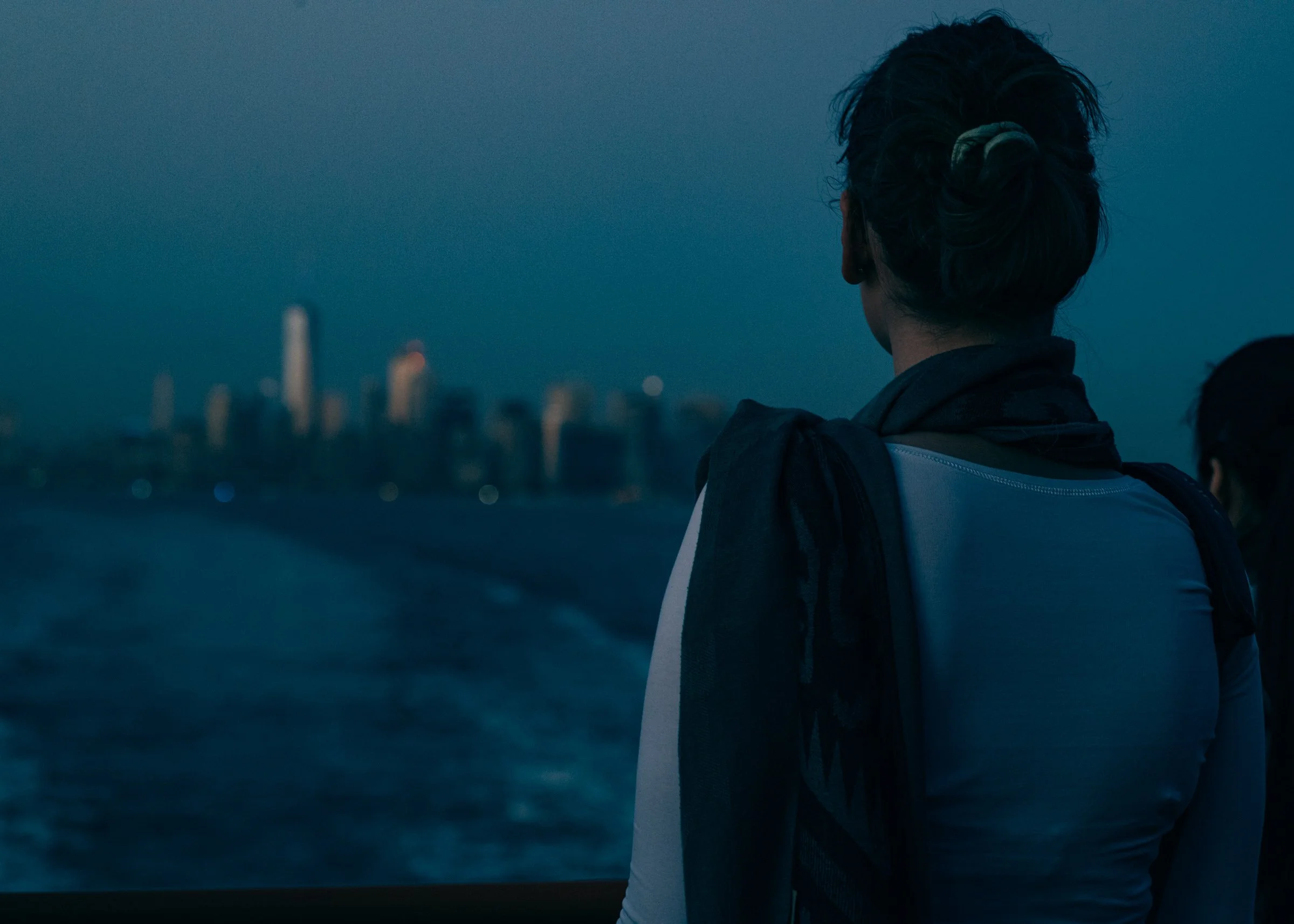 A woman with her hair in a bun and a scarf over her shoulder seen from behind at the ferry rail at dusk, watching the lit Lower Manhattan skyline recede across the boat's broad white wake.