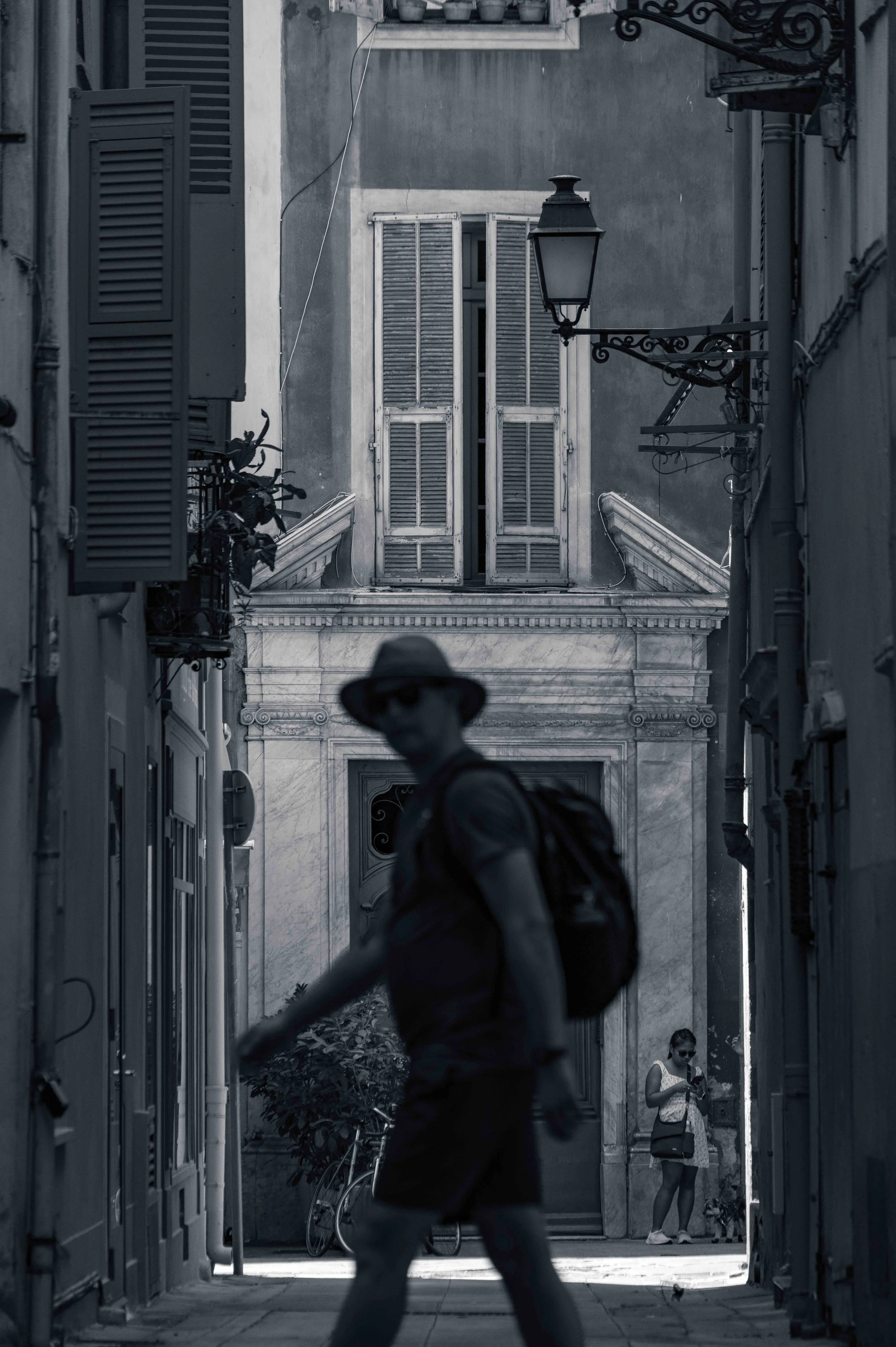 A man in a hat with a backpack walks through a narrow, deeply shaded old-town street in Nice, lit by a single strip of sunlight.