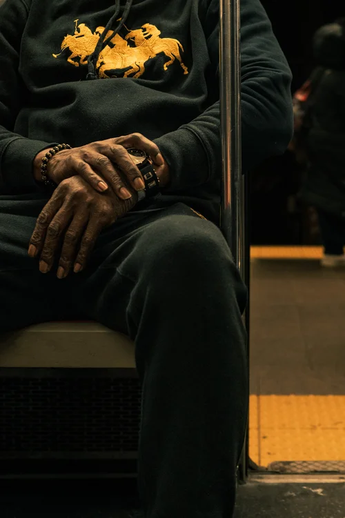 A seated figure in a dark hooded top, hands resting on their knees, shown from the shoulders down against warm low light in New York.