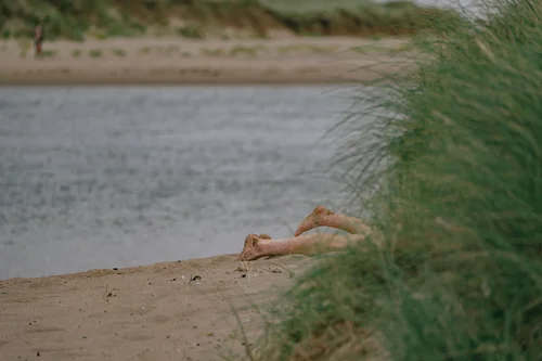 A person reclining on the sand at Malahide, Dublin, glimpsed through the dune grass with the calm estuary water beyond.