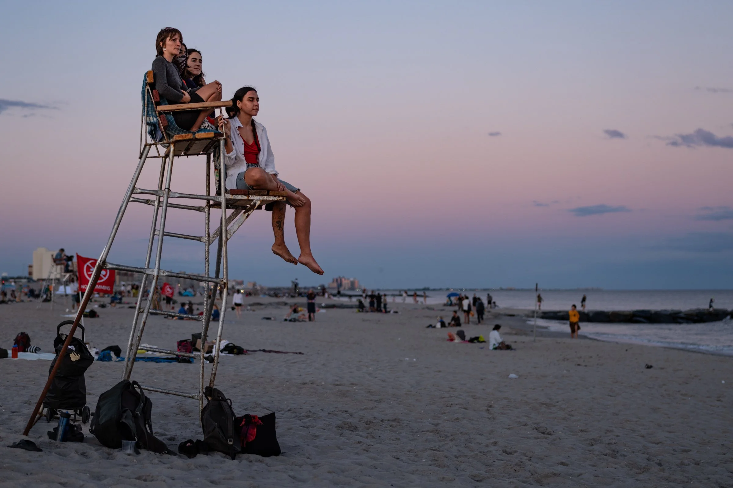 Three young women sitting on a wooden lifeguard chair on the sand at dusk at Rockaway Beach, Queens, watching a public dance, with a pink and blue sky over the ocean.