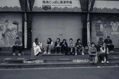 A row of people resting on the kerb before shuttered storefronts at the fish market, Tokyo, in black and white.