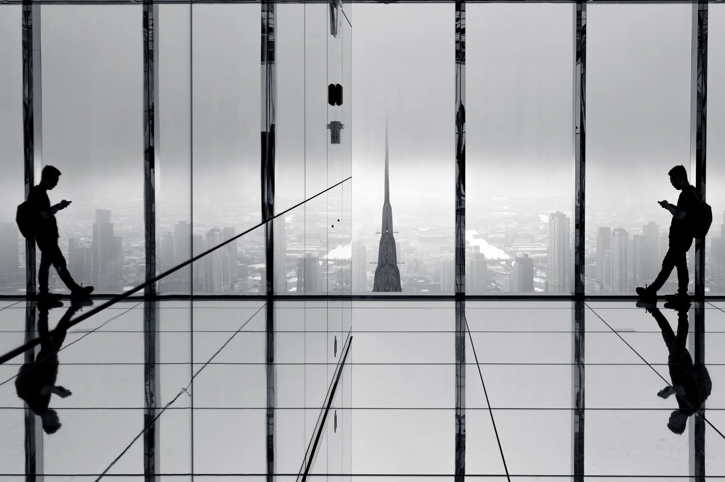 Reflection of a man in silhouette standing at floor-to-ceiling glass on a high observation deck, his reflections repeated in the mirrored floor below him, the spire of the Chrysler Building dead centre in flat white haze, in black and white.