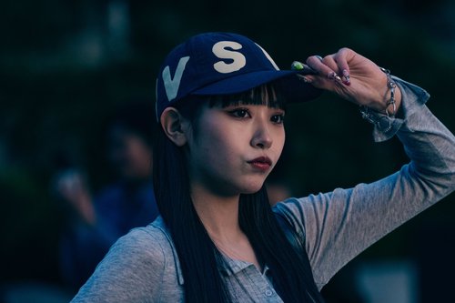 A young woman in a cap adjusting the brim, city lights blurred behind her at night, Tokyo.