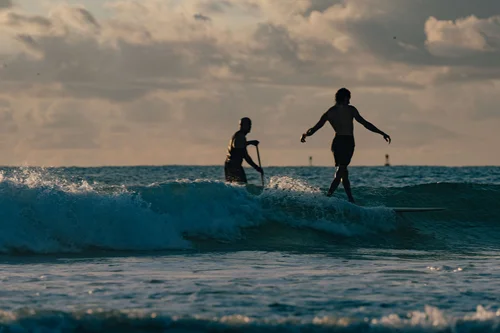 Two figures silhouetted at the shoreline at sunrise, one caught mid-movement, a wave breaking in front, Miami Beach.