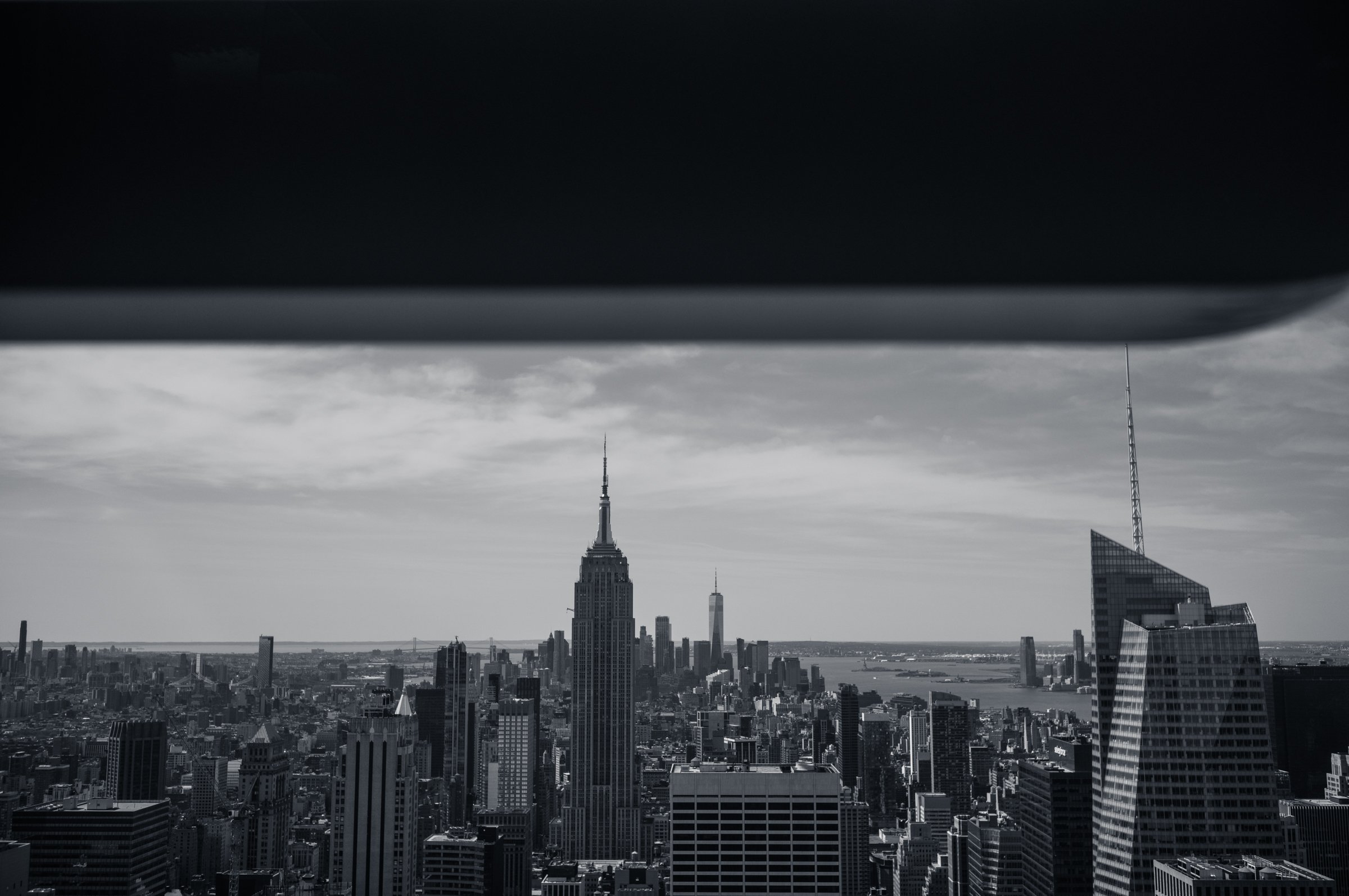 The view south from the Top of the Rock observation deck with the Empire State Building at the centre, One World Trade Center beyond it and the harbour in haze, seen from under a dark overhang, in black and white.