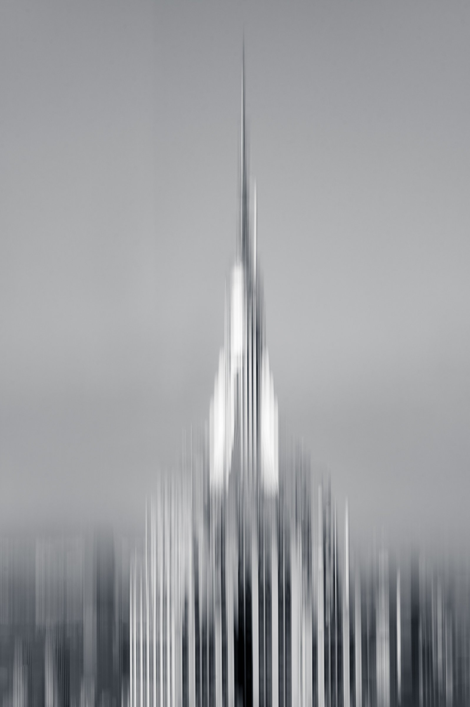 A tall spire dissolved into fine vertical streaks by a moving camera, the buildings beneath it pulled into pale ribbons, in black and white.