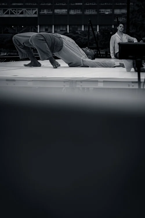 Two dancers on a low ledge, one crouched forward and one standing, with water and sky behind at Battery Park, New York, in black and white.