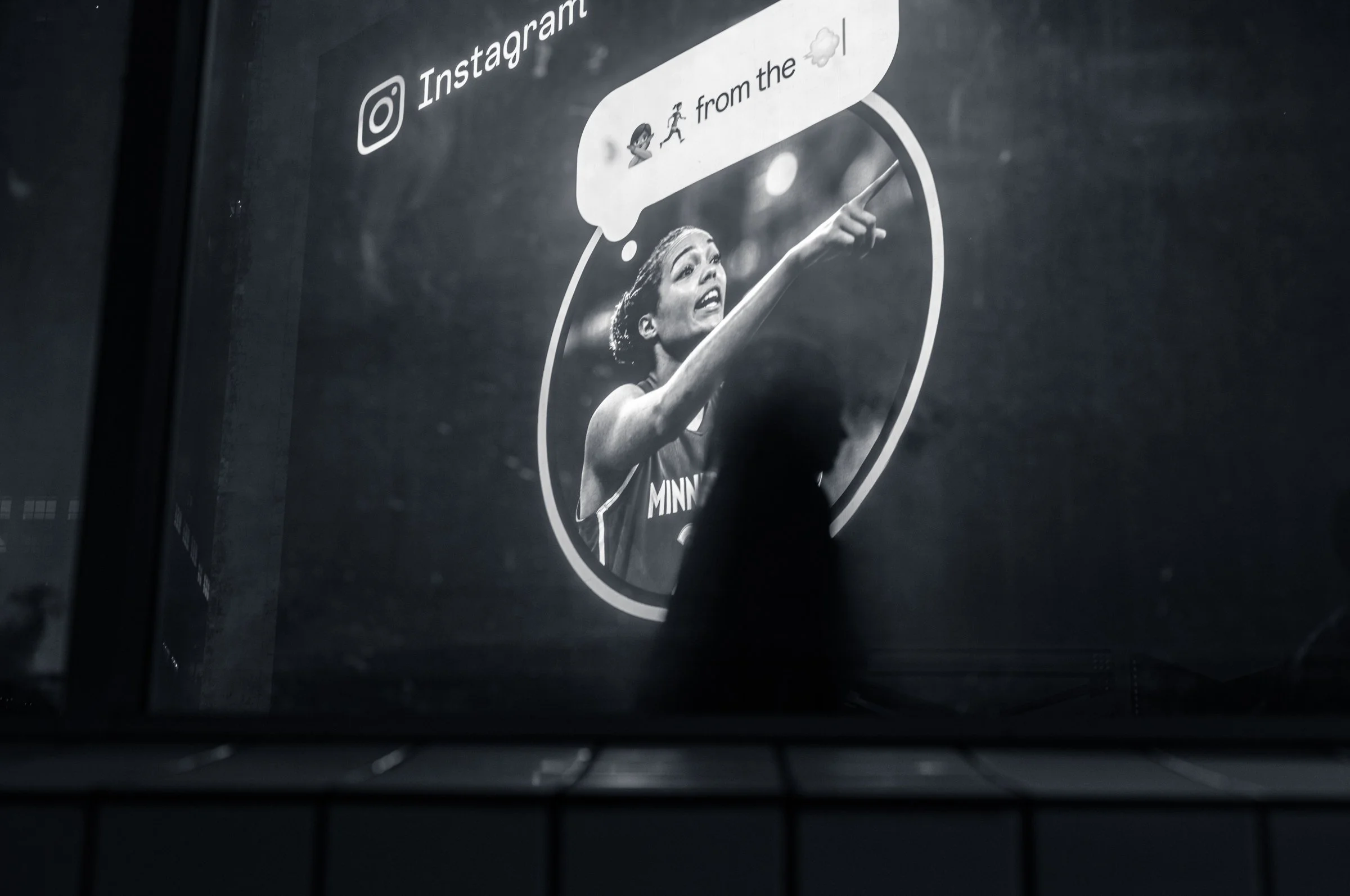 A large illuminated advertisement at night in Times Square showing a basketball player pointing, framed in a circle with a speech bubble, a person's dark silhouetted head cutting across the bottom, in black and white.