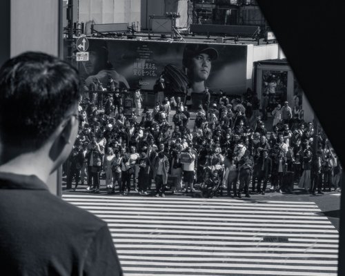 A vast crowd surging across the road at Shibuya Crossing, seen over a bystander's shoulder, Tokyo, in black and white.