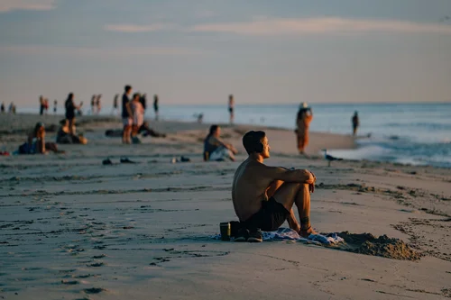 A person sitting alone on the sand in the foreground as a crowd gathers at the water's edge behind, Miami Beach.