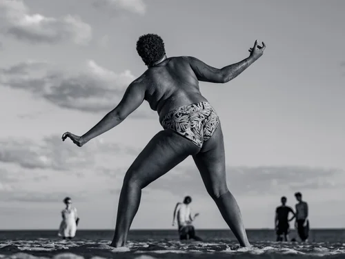 A dancer in a wide, grounded stance with arms outstretched on the sand, the sea and distant bathers behind at Rockaway Beach, New York, in black and white.